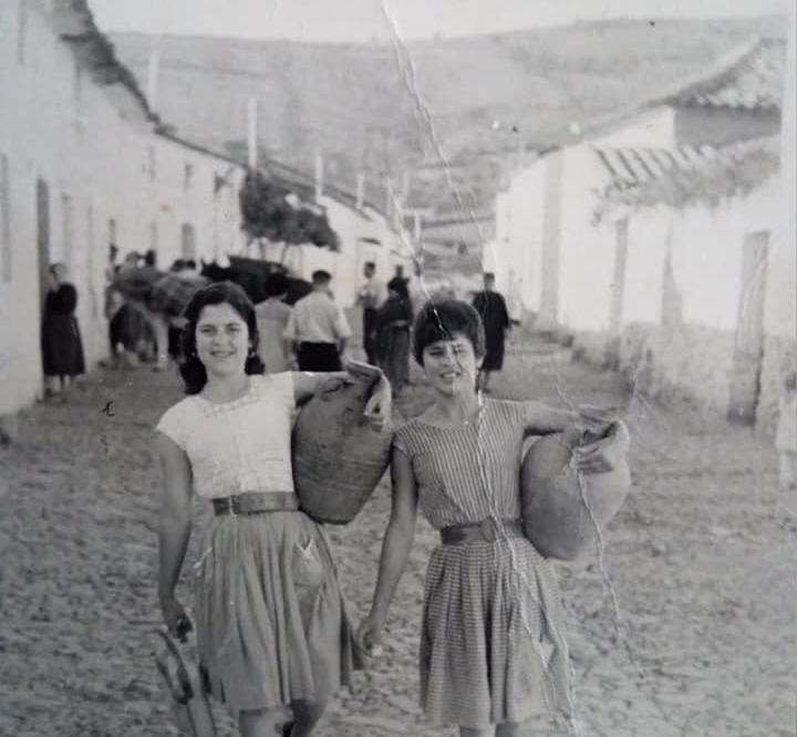Fotografía en B/N de dos jóvenes llevando cántaros de agua en una calle rural empedrada de casas blancas.